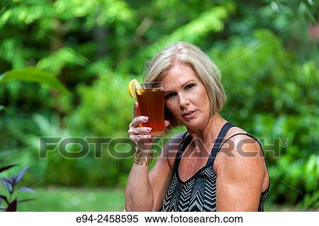 A 56 year old blond woman holding a glass of iced tea holding it to her forehead looking at the camera. View Large Photo Image Stock Photography - A 56 year old blond woman holding a glass of iced tea holding it to her forehead looking at the camera.. Fotosearch