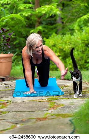 A 56 year old blond woman on a yoga mat, reaching out to pet a cat, outdoors. View Large Photo Image Stock Image - A 56 year old blond woman on a yoga mat, reaching out to pet a cat, outdoors.. Fotosearch