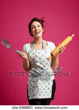 Stock Image - Funny housewife and kitchen utensils, pink background.. Fotosearch