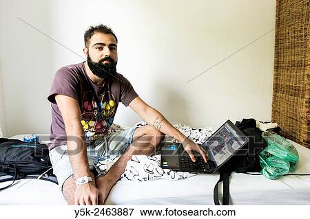 Tilburg, Netherlands. Brazilian male traveller sitting on the bed of his Airbnb guest room with his laptop computer at arms length. View Large Photo Image Stock Photo - Tilburg, Netherlands. Brazilian male traveller sitting on the bed of his Airbnb guest room with his laptop computer at arms length.. Fotosearch