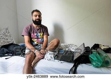 Tilburg, Netherlands. Brazilian male traveller sitting on the bed of his Airbnb guest room with his laptop computer at arms length. View Large Photo Image Stock Photo - Tilburg, Netherlands. Brazilian male traveller sitting on the bed of his Airbnb guest room with his laptop computer at arms length.. Fotosearch