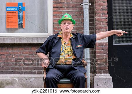 Tilburg, Netherlands. Man sitting in his newly bought chair in front of a facade in the streets and pointing towards a situation at his left. View Large Photo Image Stock Photography - Tilburg, Netherlands. Man sitting in his newly bought chair in front of a facade in the streets and pointing towards a situation at his left.. Fotosearch