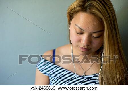 Tilburg, Netherlands. Studio portrait of a young Chinese girl with headphones, listening to the music on her smartphone, while standing in front of a grey background. View Large Photo Image Stock Photo - Tilburg, Netherlands. Studio portrait of a young Chinese girl with headphones, listening to the music on her smartphone, while standing in front of a grey background.. Fotosearch