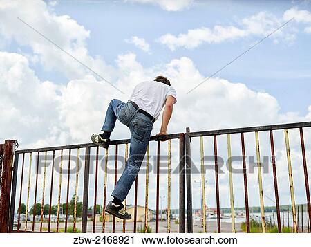Stock Image - Young adult male climbs the fence, overcast sky on background. Shot in southern Finland.. Fotosearch