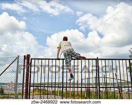 Stock Image - Young adult male climbs the fence, overcast sky on background. Shot in southern Finland.. Fotosearch
