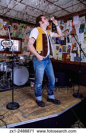 Stock Image - Beatnick looking male chugging beer onstage decorated with posters and a drum set wearing cuffed jeans.. Fotosearch