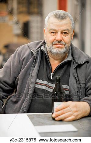 Mature caucasian man with beer bootle sitting at a table in his shop. View Large Photo Image Stock Photograph - Mature caucasian man with beer bootle sitting at a table in his shop.. Fotosearch