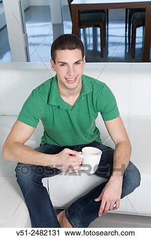 Stock Image - Portrait of a young man sitting on sofa with coffee and smiling at camera. Fotosearch