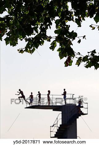 Stock Image - selected focus, focus on the tree leaves in the foreground, out of focus background - single man jumping from diving tower to waters of Lake Geneva, Paquis beach, Geneva, Switzerland. Fotosearch