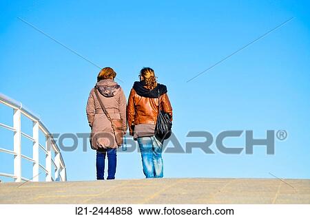 Two women back, wearing both with coat, handbag and jeans. Figures silhouetted against the blue sky, near a white railing. Barcelona, Catalonia, Spain. View Large Photo Image Stock Photo - Two women back, wearing both with coat, handbag and jeans. Figures silhouetted against the blue sky, near a white railing. Barcelona, Catalonia, Spain.. Fotosearch