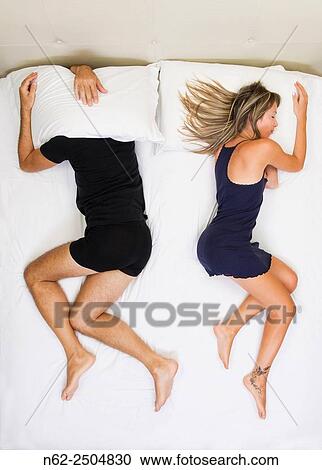 Stock Image - Unhappy couple sleeping far away from each other on bed, him has his head under the pillow, top view.. Fotosearch