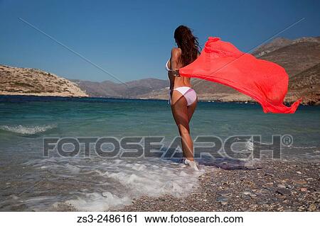 Stock Image - Woman holding an orange scarf in her hand at the beach, Amorgos, Cyclades Islands, Greek Islands, Greece, Europe.. Fotosearch