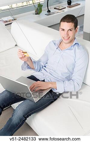 Stock Image - Young man buying in internet with credit card sitting on sofa smiling at camera. Fotosearch