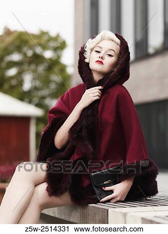 Stock Image - Fashionable blond wearing red coat and she sits on a city bench, urban scene.. Fotosearch