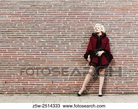 Stock Image - Fashionable blond wearing red coat, brick wall on background, urban scene.. Fotosearch
