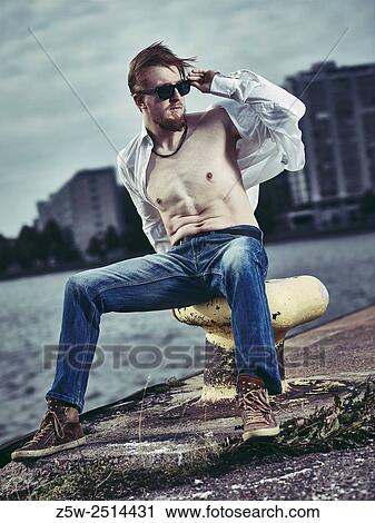 Stock Image - Handsome man wearing white shirt and jeans, windy day in harbour.. Fotosearch