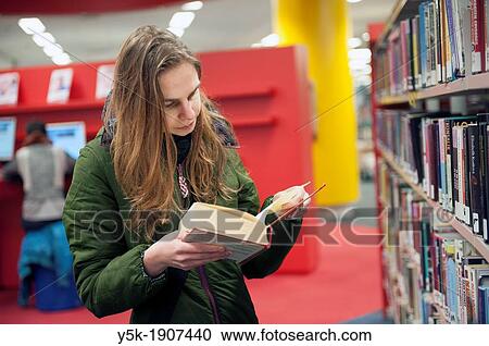 Tilburg, Netherlands. Young brunette reading a book in the public library, standing between the bookshelves with books. View Large Photo Image Stock Image - Tilburg, Netherlands. Young brunette reading a book in the public library, standing between the bookshelves with books.. Fotosearch