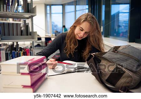 Stock Image - Tilburg, Netherlands. Young brunette studying the texts of some books in the public library, while sitting at a table.. Fotosearch