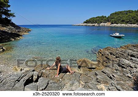 Stock Image - young woman sitting on rocks at a creek of Mrcara islet in the north-west of Lastovo island, Croatia, Southeast Europe.. Fotosearch