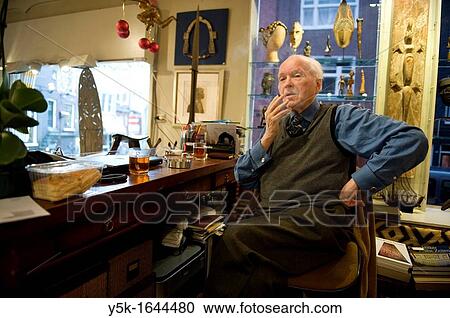 Stock Image - Amsterdam, Netherlands. An old man sitting in his chair behind his desk in his antique shop with Africa art.. Fotosearch