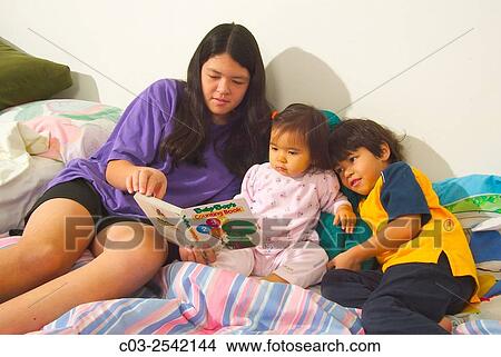 Picture - An Asian American teen baby sitter reads to her infant sister and younger brother in bed at home in Laguna Beach, CA.. Fotosearch