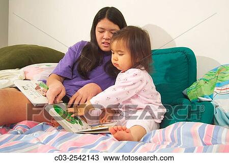 Stock Image - An Asian American teen girl reads to her infant sister in bed while babysitting in Laguna Beach, CA. MODEL RELEASE.. Fotosearch