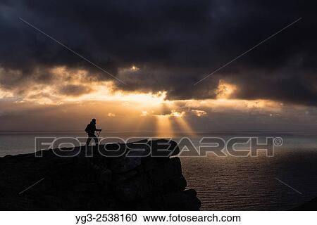 Stock Image - Female hiker watches midnight sun over sea from Nonstind mountain peak, Vestvågøy, Lofoten Islands, Norway.. Fotosearch