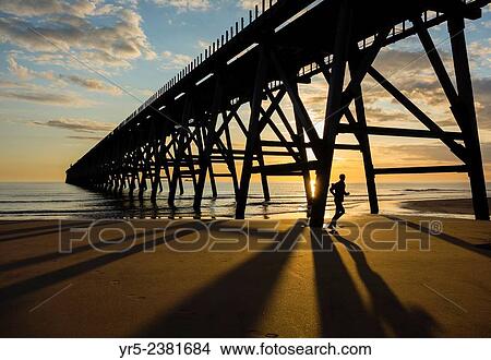 Picture - Jogger at sunrise under Steetley pier on The Headland, Hartlepool, north east England.. Fotosearch