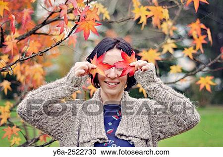 Stock Image - Smiling mid adult woman holding leaf close to her face, autumn. Fotosearch
