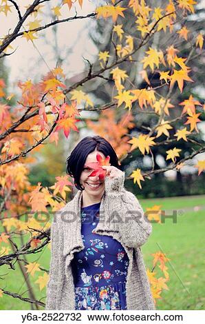 Stock Image - Smiling mid adult woman holding leaf close to her face, autumn. Fotosearch