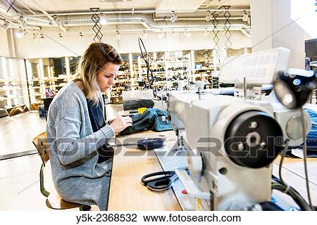 Stock Image - Tilburg, Netherlands. Young needlewoman buzzy shortening a jeans at her workstation in a fashion store for one of her customers.. Fotosearch