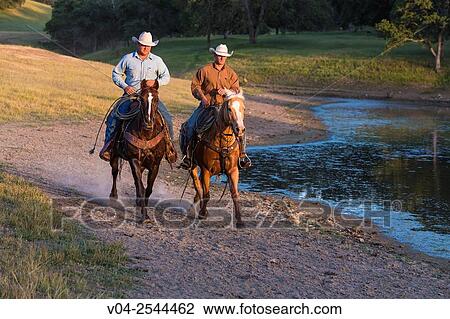 Stock Image - Two wranglers (cowboys) on horses, riding near a pond, California, USA. Fotosearch