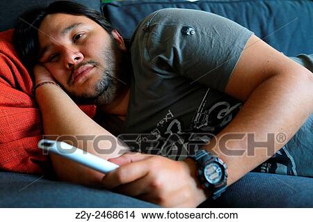 YOung man laying on a couch with TV remote on his hand. View Large Photo Image Picture - YOung man laying on a couch with TV remote on his hand.. Fotosearch