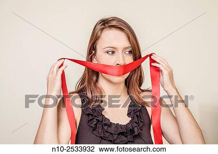Stock Image - Young woman holding a red ribbon over her mouth.. Fotosearch