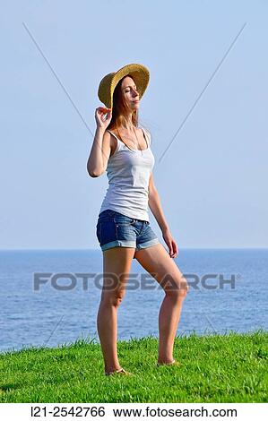 Young woman in summer clothes and straw hat standing on the grass near the sea. She is dressed with short jeans and a white blouse. View Large Photo Image Stock Photograph - Young woman in summer clothes and straw hat standing on the grass near the sea. She is dressed with short jeans and a white blouse.. Fotosearch