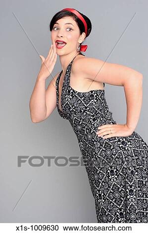 Stock Image - A 20-something attractive female in a sleeveless dress with a red ribbon in her hair has one hand on her hip and the other held to her mouth expressing surprise.. Fotosearch
