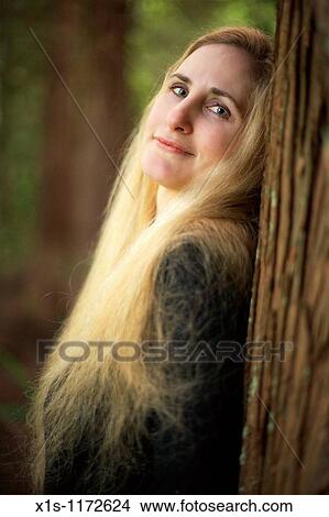 A beautiful woman with very long blonde hair leans back against a tall tree and looks toward viewer with a soft smile. View Large Photo Image Picture - A beautiful woman with very long blonde hair leans back against a tall tree and looks toward viewer with a soft smile.. Fotosearch
