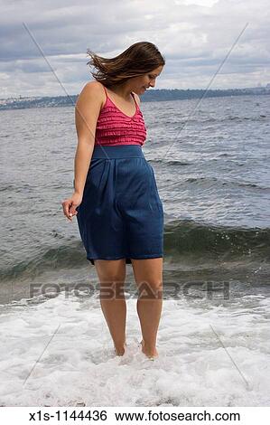 Stock Photograph - An attractive young woman wearing a skirt and top with straps walks in the waves along a beach.. Fotosearch