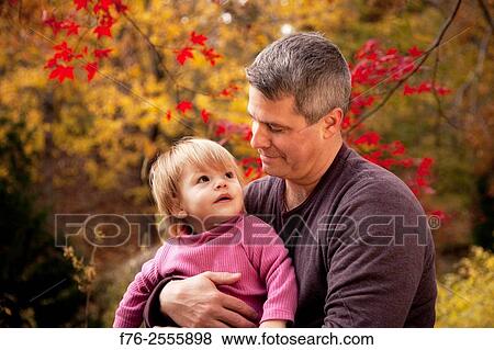 Father, in his 30s, holding his one year old daughter and holding her gaze with a loving look while sitting in their backyard in the fall with red maple leaves in the background. View Large Photo Image Stock Photo - Father, in his 30s, holding his one year old daughter and holding her gaze with a loving look while sitting in their backyard in the fall with red maple leaves in the background.. Fotosearch