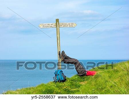 Stock Photo - Male hiker on The Cleveland Way National Trail coastal footpath resting near signpost at Skinningrove, North Yorkshire, England, United Kingdom.. Fotosearch