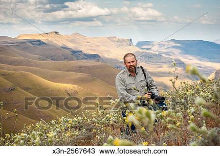 Stock Image - Man with a camera standing amongst the wildflowers with rolling hills in the background in Sani Pass, between South Africa and Lesotho.. Fotosearch