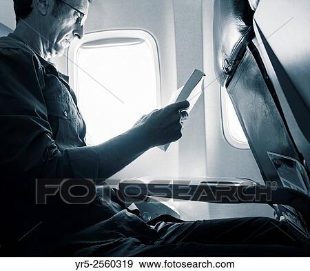 Stock Photo - Mature man reading book in window seat on airplane.. Fotosearch