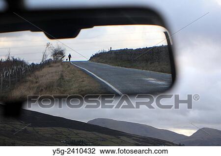 Stock Image - Person walking dog on rural road seen in car rear view mirror in wales great britain uk. Fotosearch