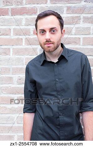 Stock Photo - Portrait of a male in his 20s wearing a black button up formal shirt with a pale brick bacground.. Fotosearch