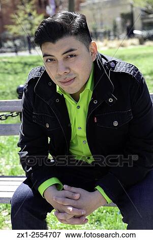 Stock Photo - Portrait of an ethnic male sitting on a bench in a park area, leaning forward with his hands clasped.. Fotosearch