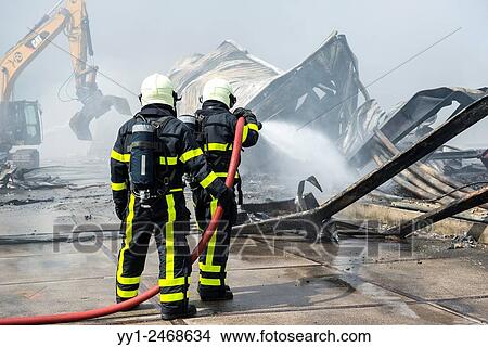 Picture - Tilburg, Netherlands. A large fire blazing inside a warehouse for plastic garden furniture at an industrial estate, with smoke nuisance as a result. Meanwhile two firemen and a Caterpillar are battling the flames from the outside.. Fotosearch