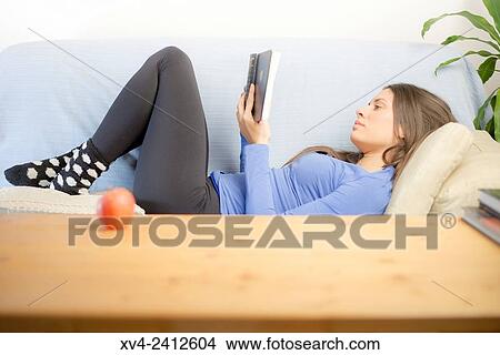 Picture - Young Woman relaxing on her sofa reading a book.. Fotosearch