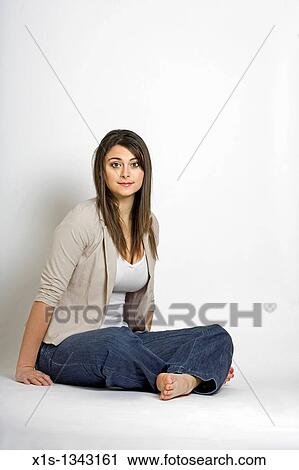 Stock Image - Young woman sits on the floor with her legs crossed, no shoes and rests her hands on the ground to support her position.. Fotosearch
