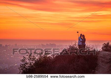 Picture - A couple using a selfie stick at sunset in Griffith Park in the Hollywood Hills overlooking Los Angeles.. Fotosearch