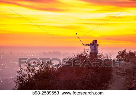Stock Image - A couple using a selfie stick at sunset in Griffith Parkin the Hollywood Hills overlooking Los Angeles.. Fotosearch